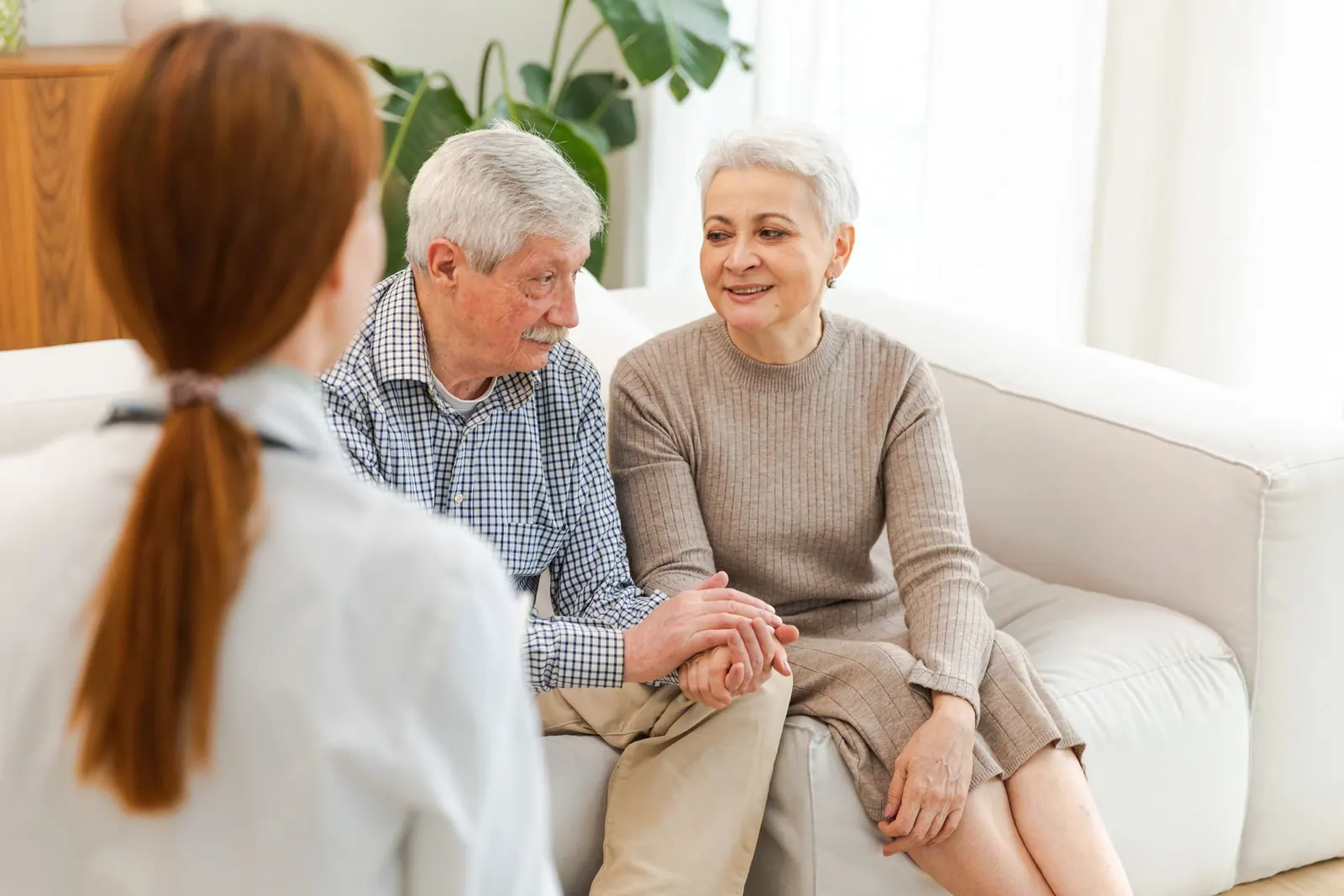 Two older adults holding hands on a couch, speaking with a caregiver in a bright living room.
