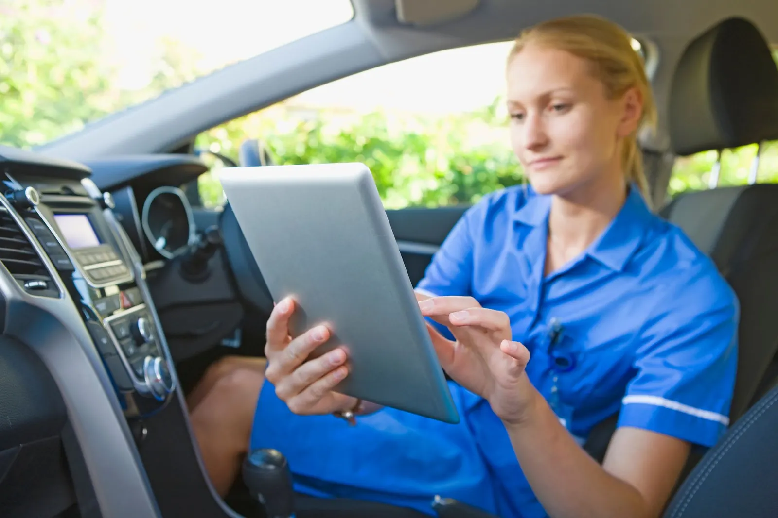 A nurse is sitting in the driver's seat of a car using a tablet.